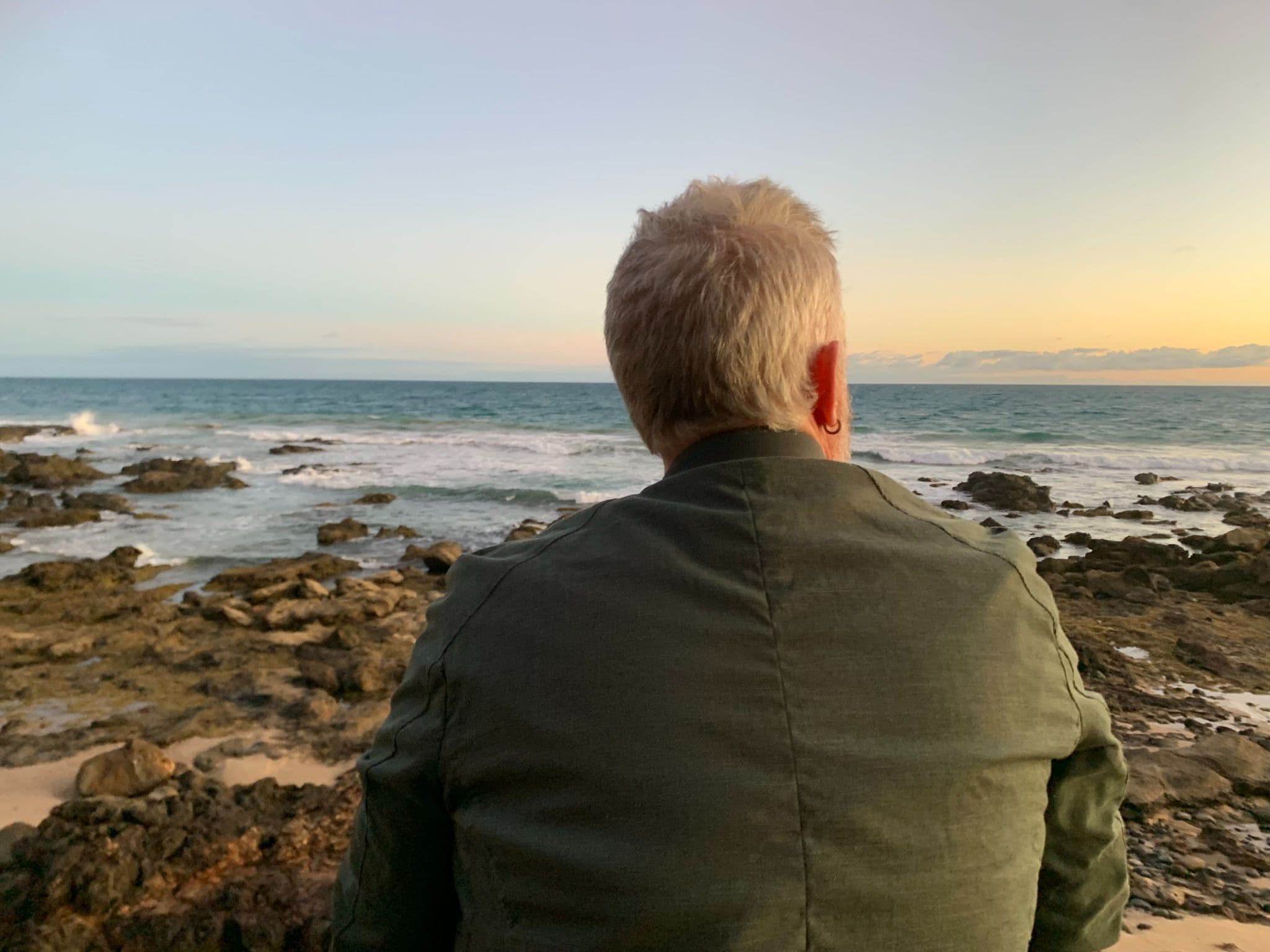 Man with grey hair standing on rocks looking out at the ocean at sunset, wearing an olive jacket