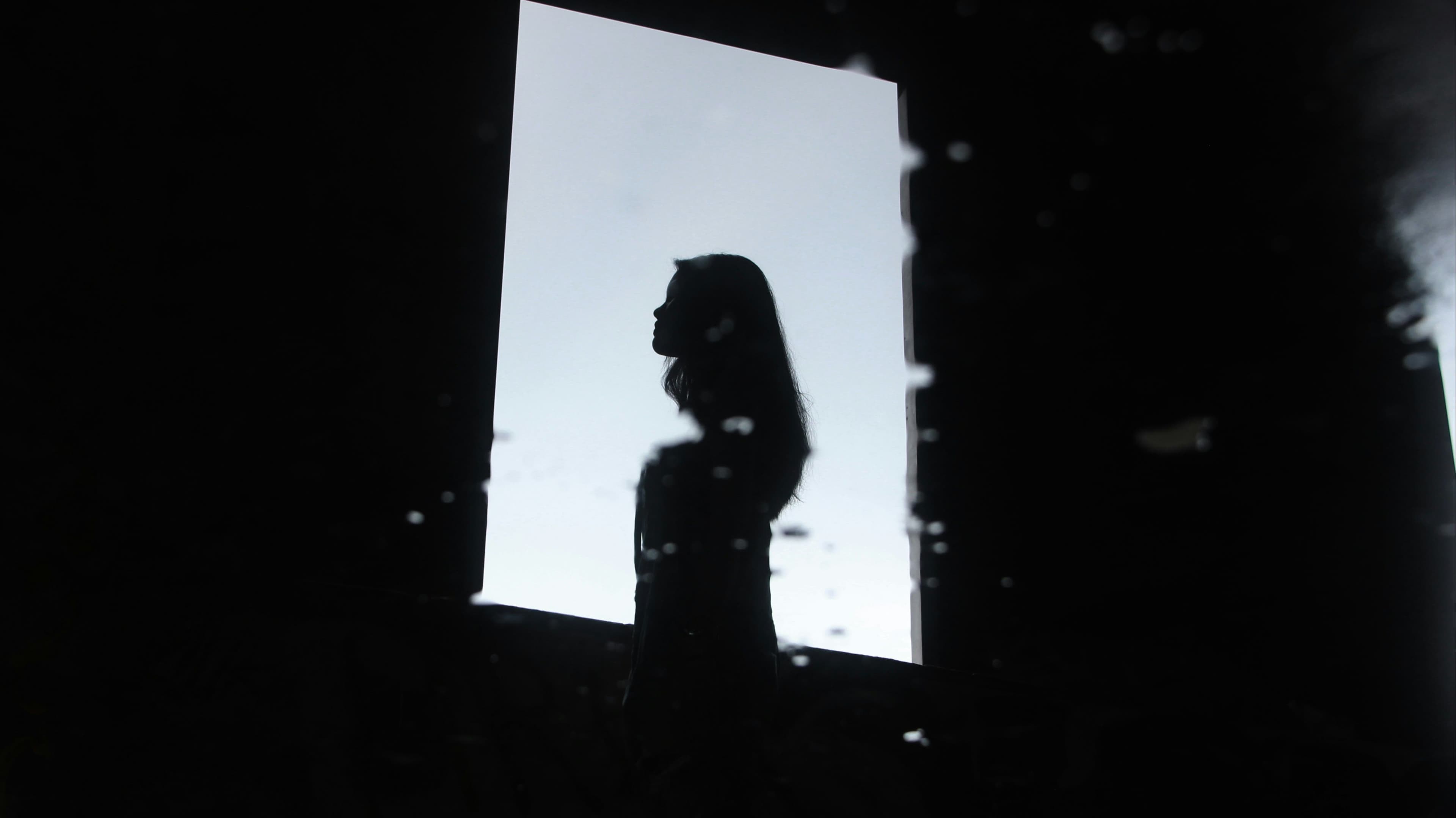 Silhouette of a woman with long hair standing before a glowing white backdrop in a dark room